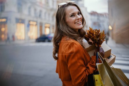 Foto de Hola octubre. mujer sonriente de moda en gabardina marrón con paquetes, bolsas de compras y hojas amarillas de otoño en la ciudad. - Imagen libre de derechos