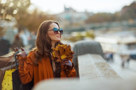 Foto de Hola noviembre. mujer elegante sonriente en gabardina naranja con bolsas de compras y hojas amarillas de otoño en la ciudad. - Imagen libre de derechos
