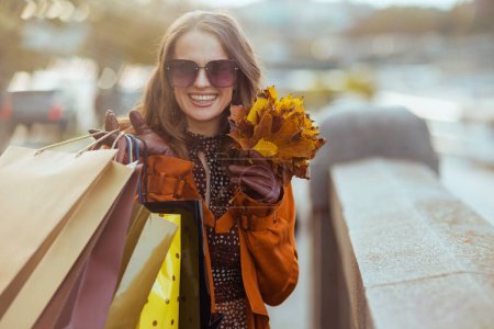 Foto de Hola otoño. mujer joven feliz en abrigo naranja con bolsas de compras y hojas amarillas de otoño en la ciudad. - Imagen libre de derechos