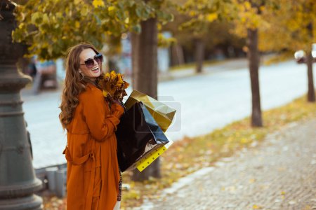 Foto de Hola otoño. mujer de moda feliz en gabardina naranja con bolsas de compras y hojas amarillas de otoño en la ciudad. - Imagen libre de derechos