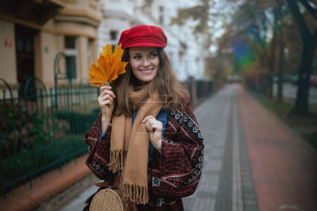 Foto de Hola otoño. sonriente mujer moderna en sombrero rojo con hojas de otoño y bufanda caminando por la ciudad. - Imagen libre de derechos