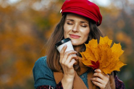 Téléchargez les photos : Bonjour l'automne. élégante femme détendue en chapeau rouge avec des feuilles d'automne, écharpe et café dans le parc de la ville. - en image libre de droit