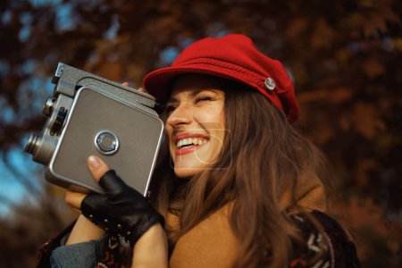 Foto de Hola otoño. feliz elegante mujer de mediana edad en sombrero rojo con cámara de vídeo retro, bufanda y guantes en el parque de la ciudad. - Imagen libre de derechos