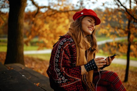 Photo for Hello autumn. happy stylish middle aged woman in red hat with scarf, smartphone and gloves listening to the music with headphones while sitting on bench in the city park. - Royalty Free Image