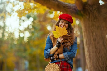 Téléchargez les photos : Bonjour l'automne. Portrait de jeune femme souriante en jeans chemise et chapeau rouge avec feuille d'automne, écharpe, gants et sac près de l'arbre dans le parc de la ville. - en image libre de droit