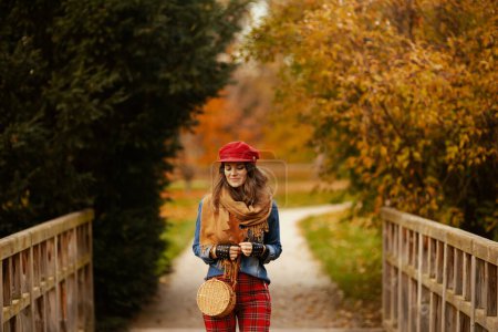 Foto de Hola otoño. pensativa joven hembra en camisa vaquera y sombrero rojo con hoja de otoño, bufanda, guantes y bolso en el parque de la ciudad. - Imagen libre de derechos