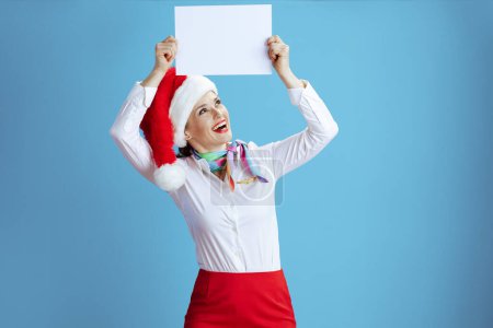 Foto de Mujer azafata elegante feliz sobre fondo azul en uniforme con Santa sombrero mostrando hoja de papel a4 en blanco. - Imagen libre de derechos