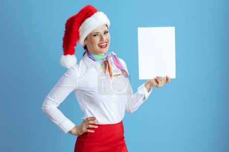 Photo for Happy elegant female air hostess isolated on blue background in uniform with Santa hat showing blank a4 paper sheet. - Royalty Free Image