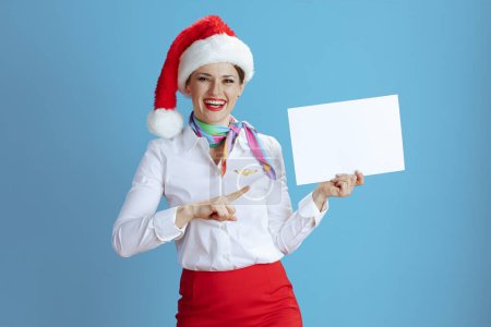 Téléchargez les photos : Souriant élégante hôtesse de l'air féminin isolé sur fond bleu en uniforme avec chapeau Santa montrant feuille de papier blanc a4. - en image libre de droit