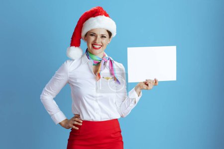 Photo for Happy elegant stewardess woman on blue background in uniform with Santa hat showing blank a4 paper sheet. - Royalty Free Image