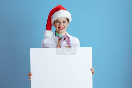 Photo for Smiling modern female flight attendant isolated on blue background in uniform with Santa hat showing blank board. - Royalty Free Image