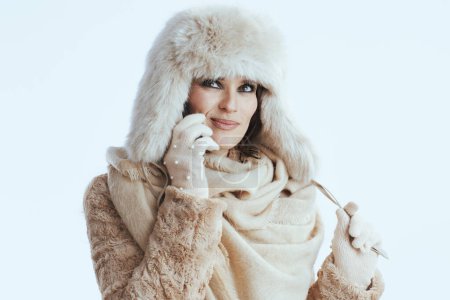 Photo for Smiling stylish middle age woman in winter coat and fur hat against white background in white gloves speaking on a smartphone. - Royalty Free Image