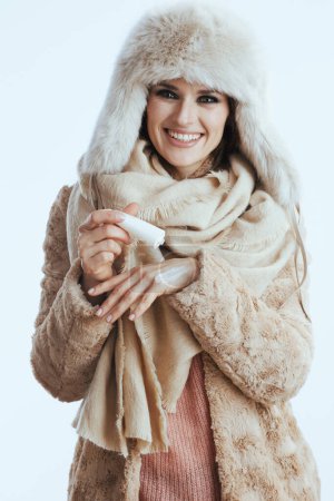 Foto de Sonriente mujer moderna de 40 años en abrigo de invierno y sombrero de piel aislado en blanco con crema de manos. - Imagen libre de derechos