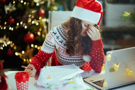 Photo for Christmas time. stressed stylish middle aged business woman in santa hat and red Christmas sweater with documents and laptop working in modern green office with Christmas tree. - Royalty Free Image