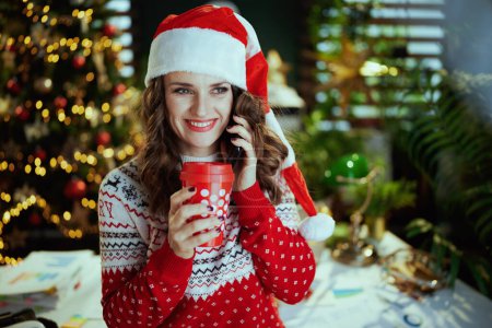 Téléchargez les photos : Noël. souriant moderne en rouge pull de Noël avec tasse de boisson chaude parler sur un smartphone en chapeau de Père Noël dans le bureau vert moderne avec arbre de Noël. - en image libre de droit