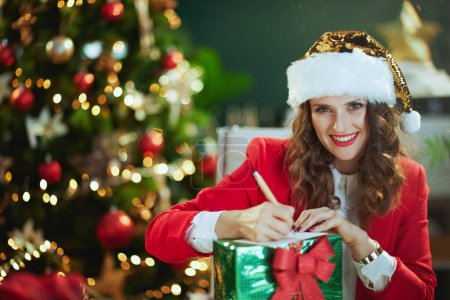 Téléchargez les photos : Noël. souriant moderne 40 ans propriétaire de petite entreprise femme en santa chapeau et veste rouge avec boîte cadeau chantant carte postale dans le bureau vert moderne avec arbre de Noël. - en image libre de droit