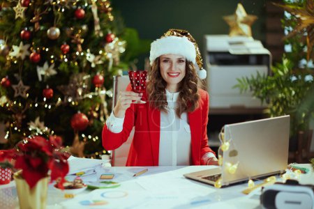 Foto de En Navidad. Retrato de mujer de negocios con estilo sonriente de 40 años en sombrero de santa y chaqueta roja con portátil y vino en la oficina verde moderna con árbol de Navidad. - Imagen libre de derechos