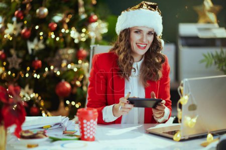 Foto de En Navidad. Retrato de feliz estilo en chaqueta roja con el ordenador portátil usando aplicación de teléfono inteligente y trabajando en sombrero de santa en la oficina verde moderna con árbol de Navidad. - Imagen libre de derechos
