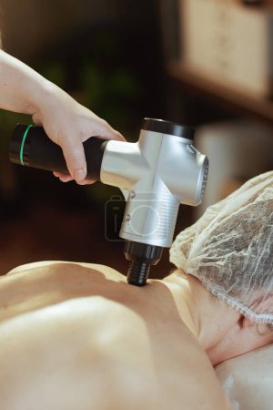 Téléchargez les photos : Temps de santé. Gros plan sur thérapeute de massage médical dans le salon de spa avec pistolet de massage massant le cou des clients. - en image libre de droit