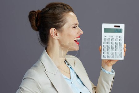 Téléchargez les photos : Souriant 40 ans propriétaire de petite entreprise femme dans un costume d'affaires léger avec calculatrice sur fond gris. - en image libre de droit