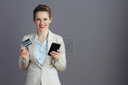 Photo for Happy young business woman in a light business suit with smartphone and credit card isolated on grey. - Royalty Free Image