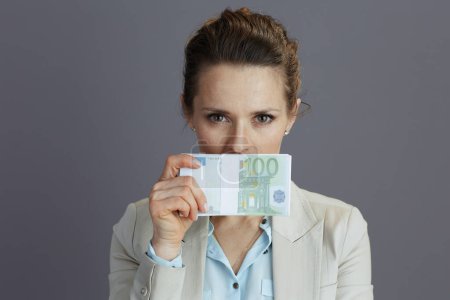 Foto de Elegante 40 años de edad, trabajadora en un traje de negocios ligero con paquetes de dinero euros que muestran no hablan mal gesto aislado sobre fondo gris. - Imagen libre de derechos