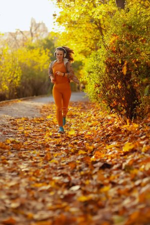 Téléchargez les photos : Bonjour l'automne. Portrait complet d'une élégante femme vêtue de vêtements de fitness dans le parc avec écouteurs en marche. - en image libre de droit