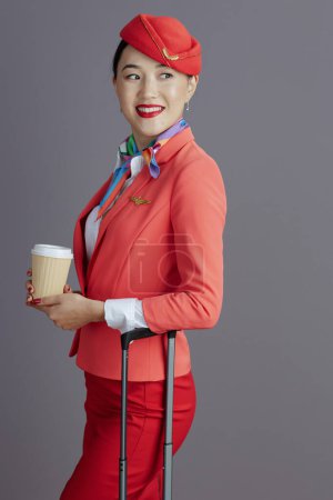 Photo for Smiling stylish asian female flight attendant in red skirt, jacket and hat uniform with coffee cup and trolley bag looking aside isolated on gray background. - Royalty Free Image