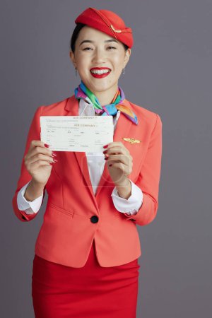 Téléchargez les photos : Heureuse hôtesse élégante asiatique femme en jupe rouge, veste et chapeau uniforme avec billets d'avion sur fond gris. - en image libre de droit