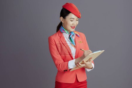 Foto de Sonriente elegante asiática azafata de vuelo en falda roja, chaqueta y sombrero uniforme con portapapeles aislado en gris. - Imagen libre de derechos