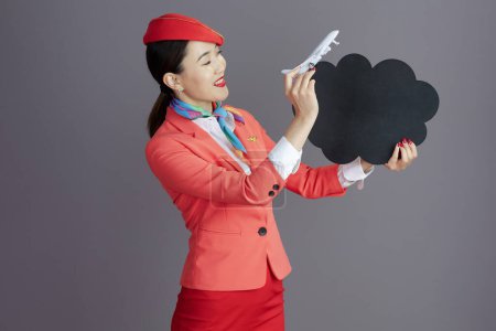 Téléchargez les photos : Heureux élégant agent de bord asiatique femme en jupe rouge, veste et chapeau uniforme avec un petit avion montrant tableau de forme de nuage vide isolé sur fond gris. - en image libre de droit