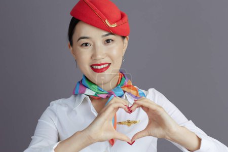 Photo for Smiling modern asian female stewardess in red skirt and hat uniform showing heart shaped hands isolated on gray background. - Royalty Free Image