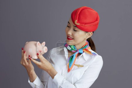 Photo for Smiling elegant air hostess asian woman in red skirt and hat uniform with piggy bank against gray background. - Royalty Free Image