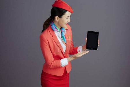 Photo for Smiling stylish asian female stewardess in red skirt, jacket and hat uniform digital tablet blank screen against gray background. - Royalty Free Image