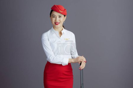 Foto de Sonriente elegante asiático azafata en rojo falda y sombrero uniforme con viaje bolsa aislado en gris fondo. - Imagen libre de derechos