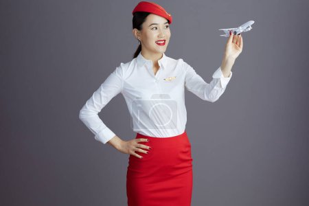 Foto de Feliz elegante azafata asiática mujer en falda roja y sombrero uniforme con un poco de avión aislado en gris. - Imagen libre de derechos