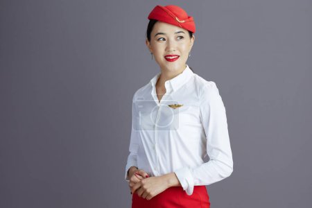 Photo for Happy elegant asian female flight attendant in red skirt and hat uniform looking at copy space isolated on grey. - Royalty Free Image
