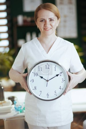 Photo for Healthcare time. happy female medical massage therapist in massage cabinet with clock. - Royalty Free Image