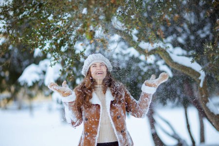 Photo for Happy modern 40 years old woman in brown hat and scarf in sheepskin coat with mittens catching snow outside in the city in winter. - Royalty Free Image
