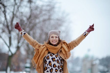 Téléchargez les photos : Femme d'âge moyen moderne souriante en chapeau marron et écharpe en manteau de chameau avec des gants se réjouissant à l'extérieur de la ville en hiver. - en image libre de droit