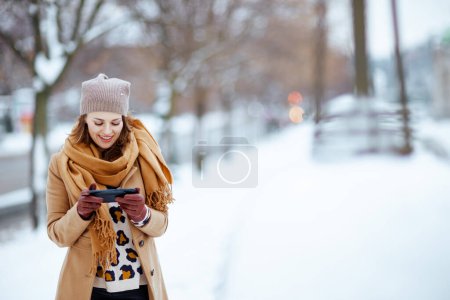 Foto de Smiling stylish woman in brown hat and scarf in camel coat with gloves sending text message using smartphone outside in the city in winter. - Imagen libre de derechos