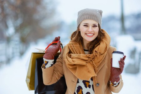 Photo for Happy stylish female in brown hat and scarf in camel coat with gloves, shopping bags and cup of soy latte outside in the city in winter. - Royalty Free Image