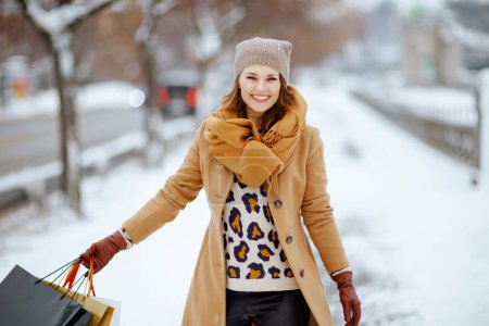Foto de Sonriente mujer moderna en sombrero marrón y bufanda en abrigo de camello con guantes y bolsas de compras en el exterior de la ciudad en invierno. - Imagen libre de derechos