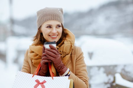 Téléchargez les photos : Heureuse élégante femme d'âge moyen en chapeau marron et écharpe en manteau de chameau avec gants, sacs à provisions et tasse de latte de soja à l'extérieur en ville en hiver. - en image libre de droit