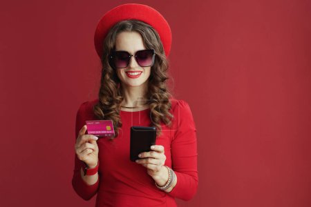 Téléchargez les photos : Joyeux Valentin. Femme élégante souriante avec de longs cheveux ondulés en robe rouge et béret sur fond rouge avec carte de crédit et smartphone acheter en ligne. - en image libre de droit