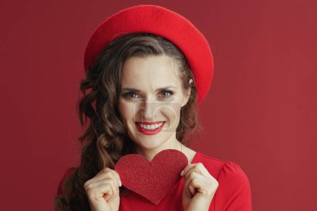 Téléchargez les photos : Joyeux Valentin. Portrait de femme élégante et souriante en robe rouge et béret isolé sur fond rouge avec cœur rouge. - en image libre de droit
