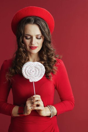 Foto de Feliz San Valentín. mujer moderna de 40 años en vestido rojo y boina aislada sobre fondo rojo con dulces en palo y pelo largo ondulado. - Imagen libre de derechos