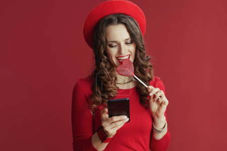 Photo for Happy Valentine. happy modern female with long wavy hair in red dress and beret against red background with heart shaped candy on stick sending text message using smartphone. - Royalty Free Image