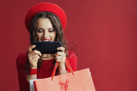 Photo for Happy Valentine. happy modern 40 years old woman in red dress and beret isolated on red background with shopping bags using smartphone applications and making online purchases on e-commerce website. - Royalty Free Image