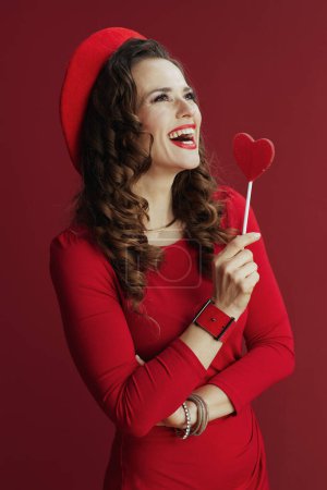 Foto de Feliz San Valentín. sonriente mujer elegante en vestido rojo y boina con corazón en forma de caramelo en palo aislado en rojo. - Imagen libre de derechos